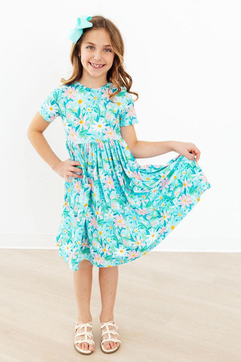 A young girl with a turquoise hair bow is wearing a short-sleeve, knee-length dress with a colorful floral and leaf print on a turquoise background. She is standing in a room with a white wall and light wood floor, holding out one side of the dress to show its flow.