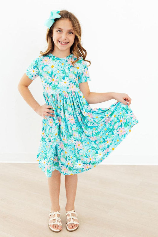 A young girl with a turquoise hair bow is wearing a short-sleeve, knee-length dress with a colorful floral and leaf print on a turquoise background. She is standing in a room with a white wall and light wood floor, holding out one side of the dress to show its flow.