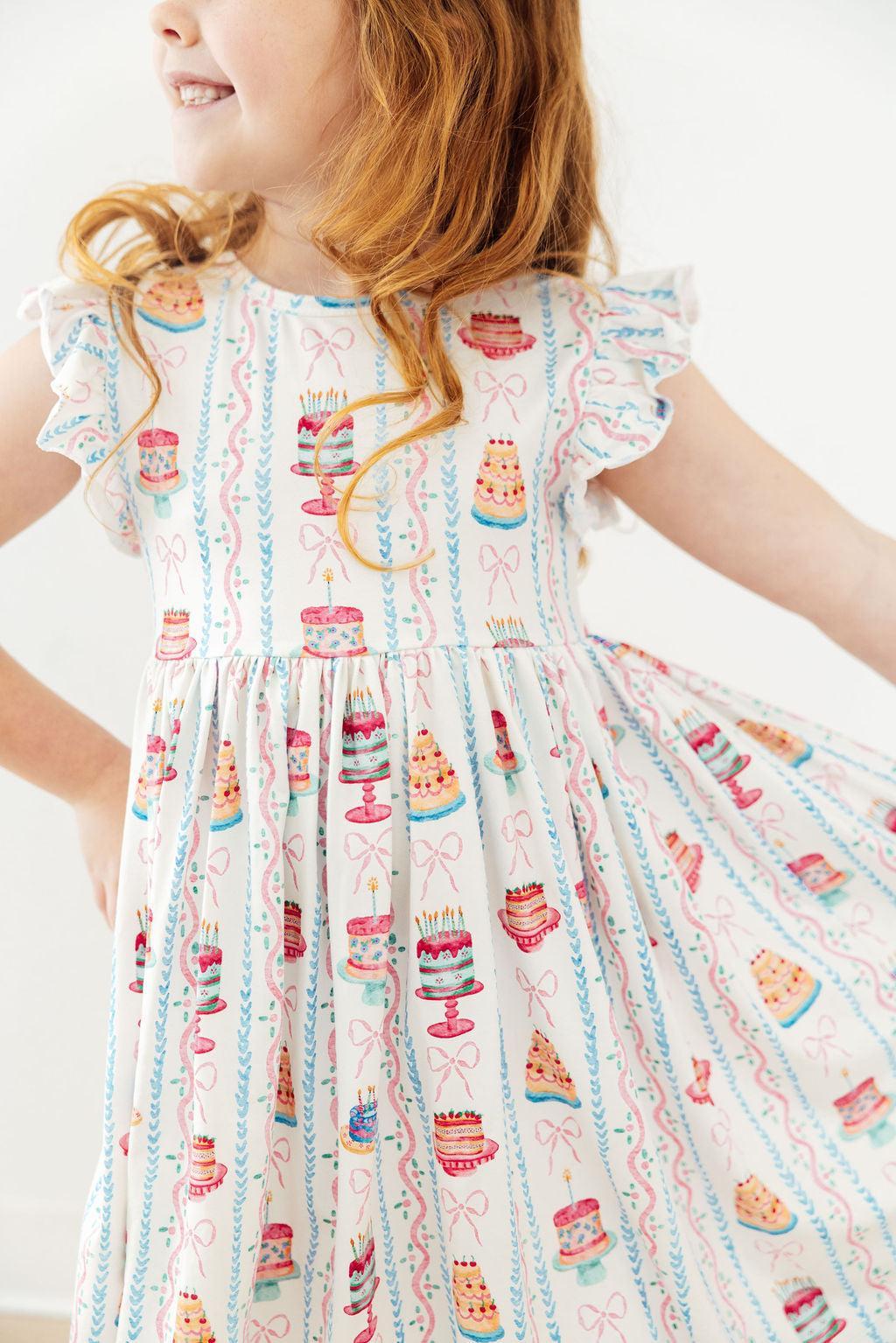 A young girl wearing a white ruffled-sleeve dress with a colorful print of cakes, candles, and bows, shown from the shoulders to the knees against a plain light background.