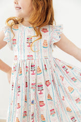 A young girl wearing a white ruffled-sleeve dress with a colorful print of cakes, candles, and bows, shown from the shoulders to the knees against a plain light background.