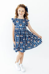 A young girl with curly light brown hair wearing a navy blue and white gingham dress with a pattern of cartoon tiger faces, paired with white sneakers and a navy hair bow, standing against a plain white background.