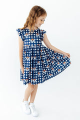 A young girl with light brown hair is wearing a blue and white gingham dress with cartoon dog prints, posing and twirling to show off the skirt. She is wearing white sneakers against a plain white background.