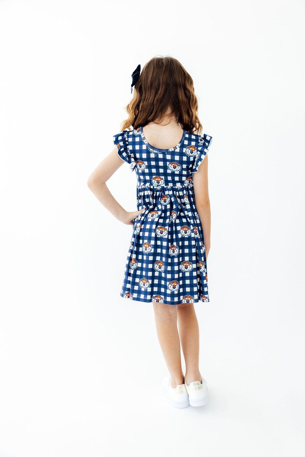 A young girl with wavy brown hair wearing a blue and white gingham dress with a teddy bear print, shown from the back against a plain white background.