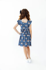A young girl with wavy brown hair wearing a blue and white gingham dress with a teddy bear print, shown from the back against a plain white background.