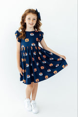 A young girl with wavy light brown hair wearing a navy blue dress with an orange and white cartoon bear pattern, white sneakers, and a matching navy hair bow, standing against a plain white background.