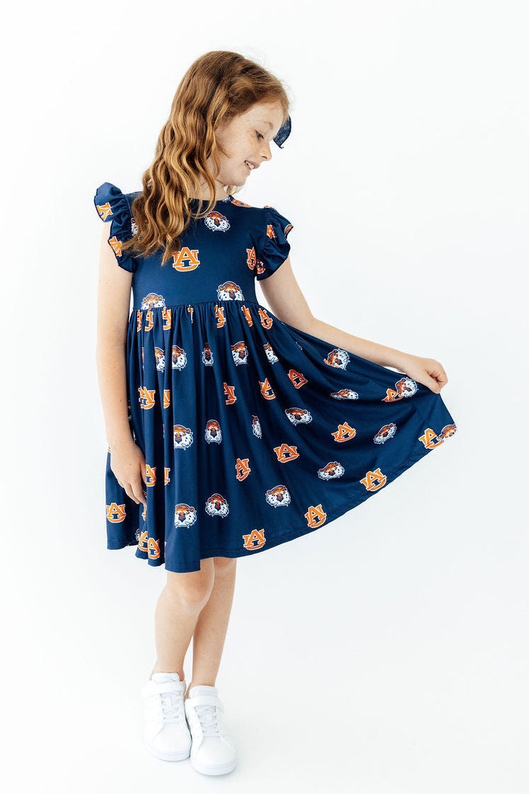 A young girl with long light brown hair is wearing a navy blue dress with a playful pattern of cartoon animals in orange, white, and blue. She is holding out the skirt to show its volume and twirling, wearing white sneakers against a clean white background.