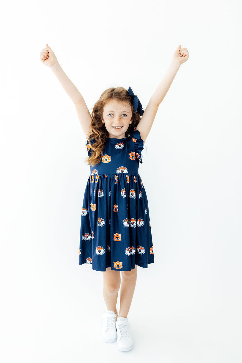 A young girl with long wavy brown hair stands with her arms raised in the air, wearing a navy blue sleeveless dress with a playful cartoon face pattern in orange, white, and yellow. She is also wearing a matching navy blue hair bow and white sneakers, set against a plain white studio background.
