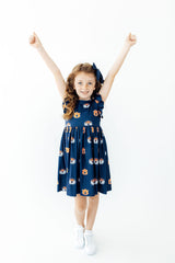 A young girl with long wavy brown hair stands with her arms raised in the air, wearing a navy blue sleeveless dress with a playful cartoon face pattern in orange, white, and yellow. She is also wearing a matching navy blue hair bow and white sneakers, set against a plain white studio background.