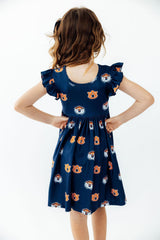 A young girl viewed from behind wearing a navy blue dress with an all-over Auburn Tigers logo print, featuring ruffled short sleeves and a gathered skirt, set against a plain white background.