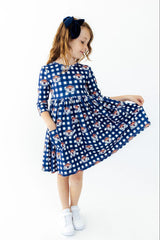 A young girl with light brown hair in a navy blue bow headband is wearing a blue and white gingham dress with a repeating pattern of cartoon dogs. She is holding out the skirt of the dress to show its volume, standing against a plain white background.