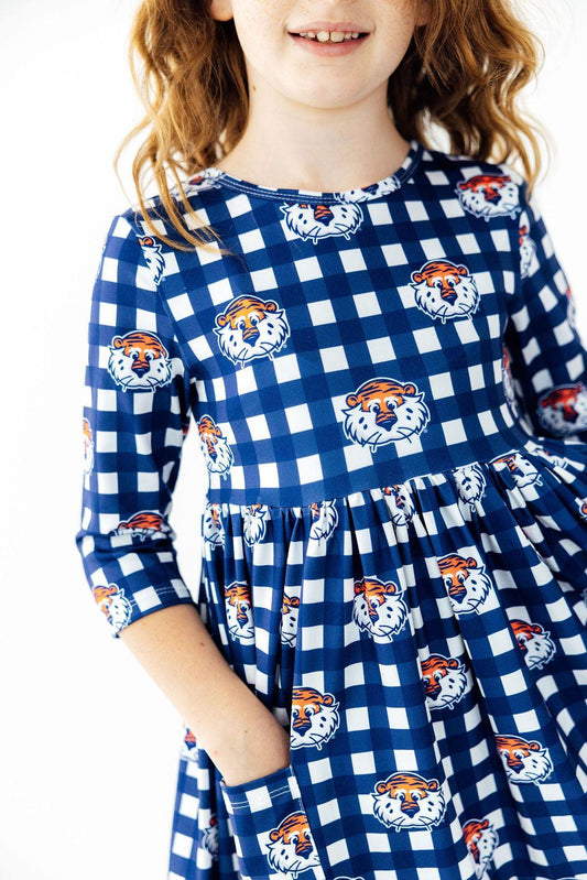 A young girl wearing a blue and white gingham dress with orange and white tiger mascot prints, shown from the shoulders down against a plain white background.