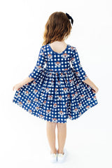 A young girl viewed from behind wearing a blue and white gingham dress with a repeating cartoon tiger face pattern, standing against a plain white background.