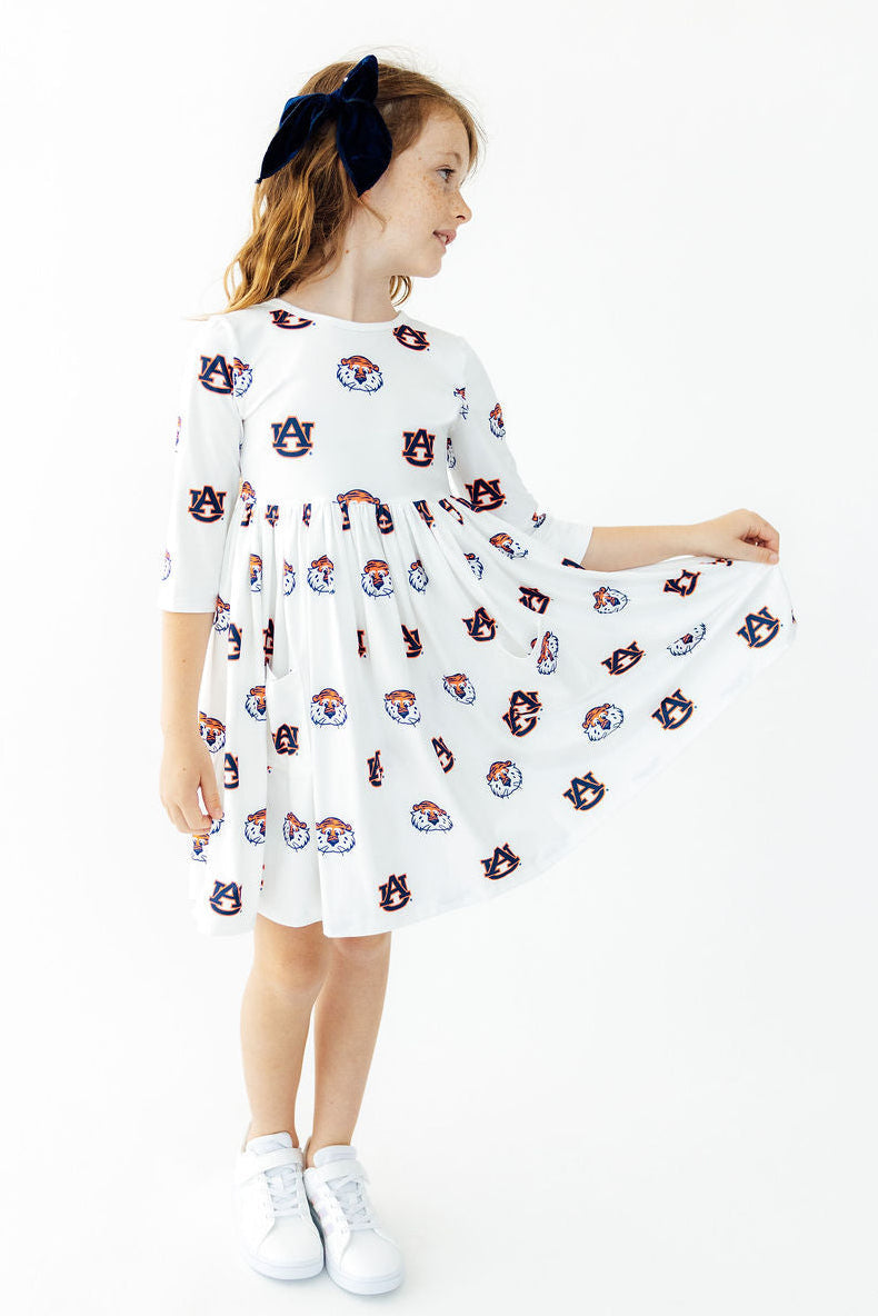 A young girl wearing a white three-quarter sleeve dress with an all-over Auburn Tigers mascot print, holding out the skirt to show its flow. She is standing against a clean white background.