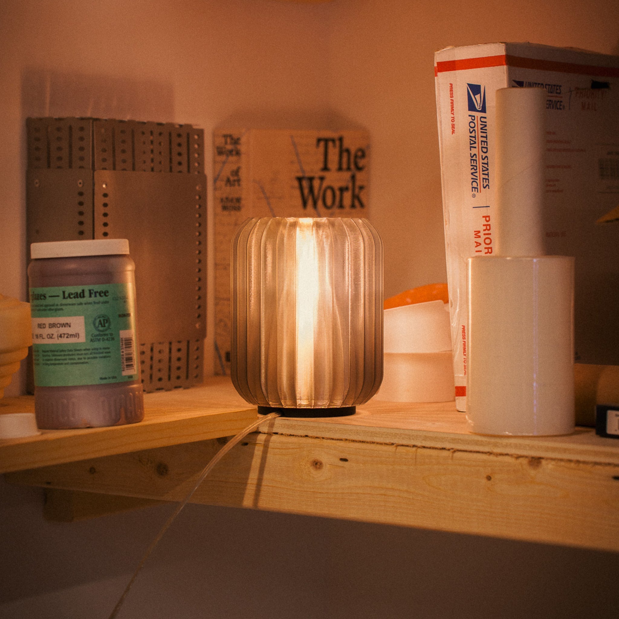 Lamp on a shelf with various items including a box labeled 'The Work' and a container of lead-free material.