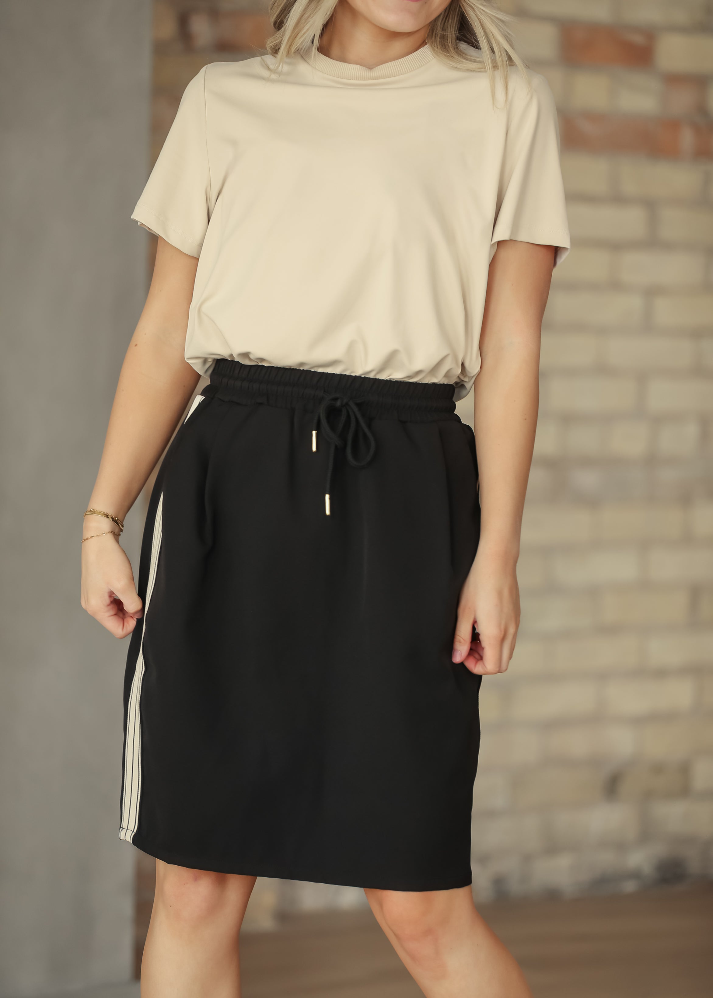 Person wearing a beige top and black skirt with white stripes against a brick wall.