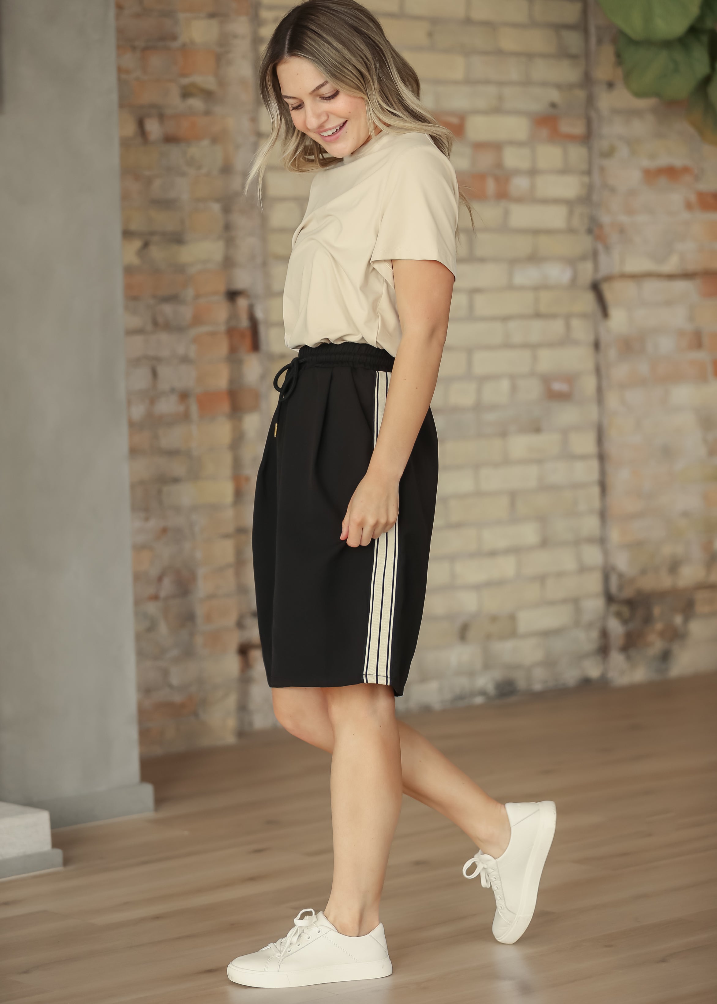 Woman wearing a beige top and black shorts with white stripes, standing against a brick wall.
