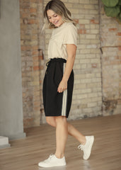 Woman wearing a beige top and black shorts with white stripes, standing against a brick wall.