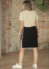 Woman wearing a beige top and black skirt with white stripes, standing against a brick wall.