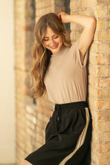 Woman leaning against a stone wall wearing a beige top and black pants with a stripe.