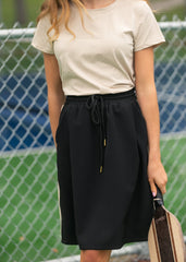 Person wearing a beige top and black skirt holding a tennis racket in front of a chain-link fence.