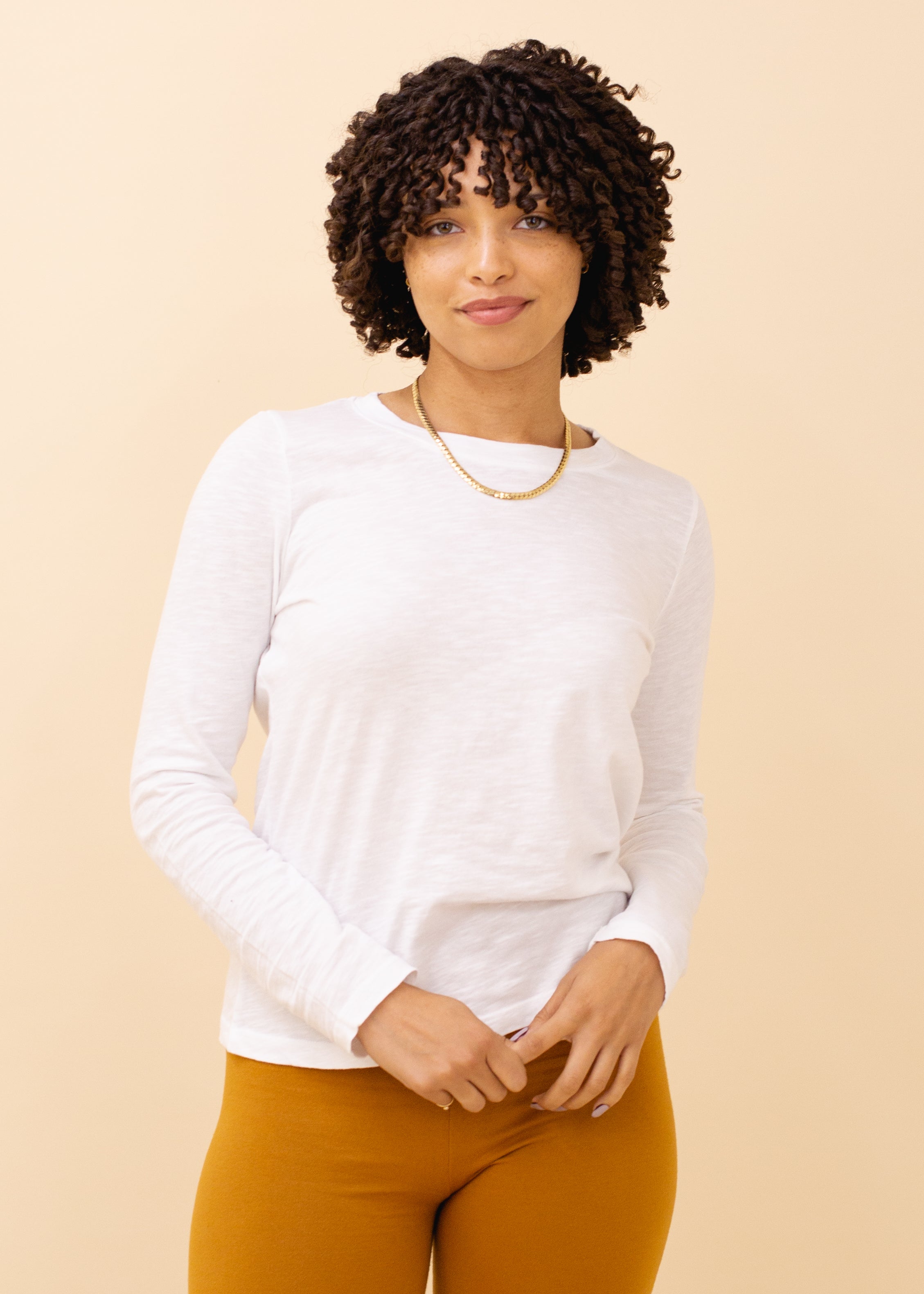 Woman wearing a white long-sleeve shirt and mustard pants against a beige background