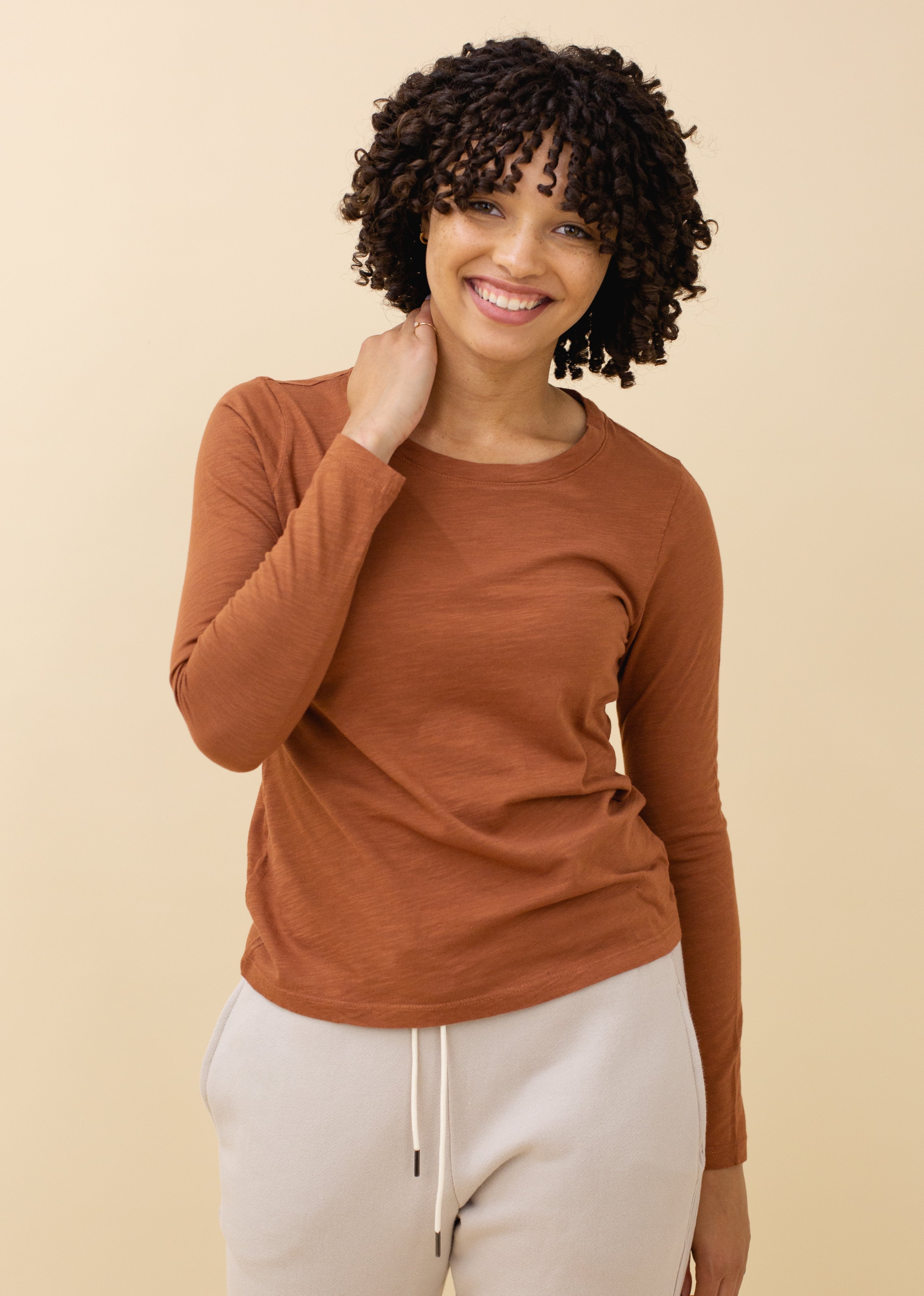 Woman wearing a brown long-sleeve top and white pants against a beige background