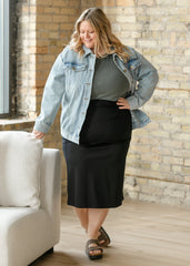 Woman wearing a denim jacket, green top, and black skirt in a room with a stone wall.