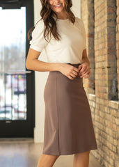 Woman wearing a white top and brown skirt standing against a brick wall.