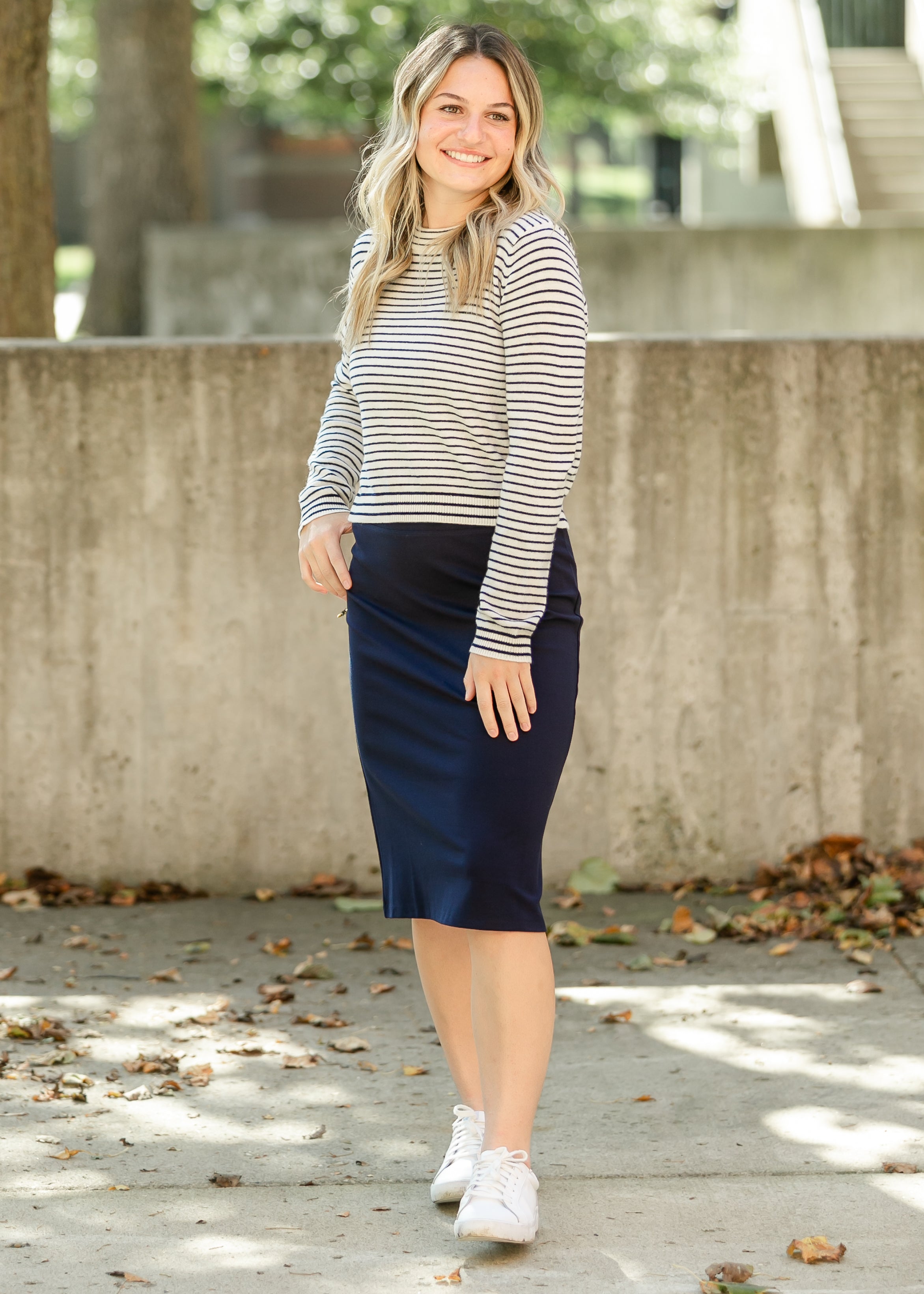 Woman wearing a striped sweater and navy skirt standing outdoors with a concrete wall and trees in the background.