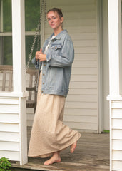 Woman wearing a denim jacket and long beige skirt standing on a porch.