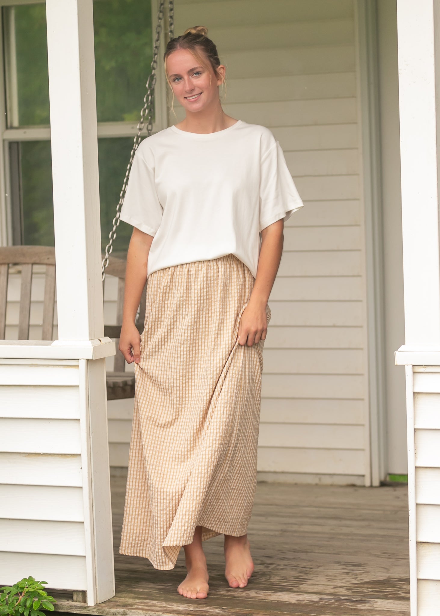 Woman standing on a wooden porch wearing a white top and beige checkered skirt.