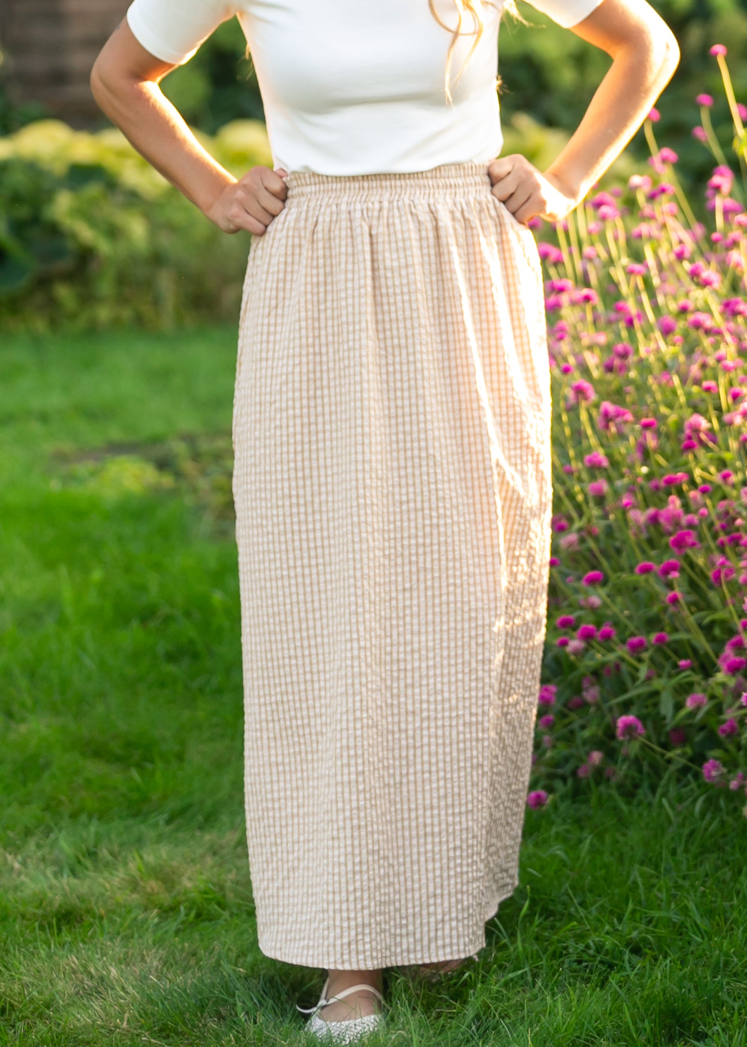 Person wearing a long beige skirt standing in a garden with green grass and pink flowers.