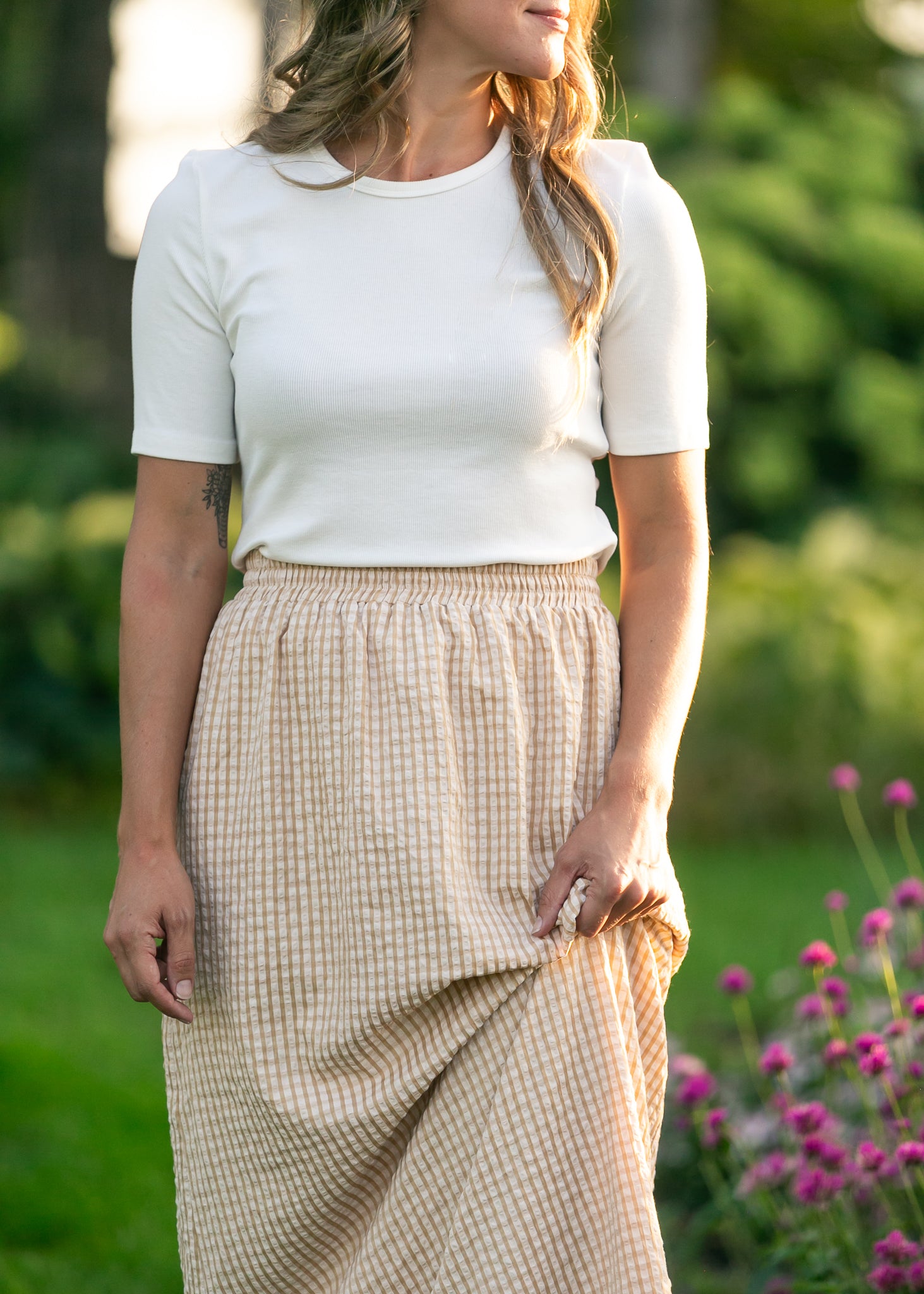 Woman wearing a white top and beige checkered skirt standing outdoors with greenery and flowers in the background