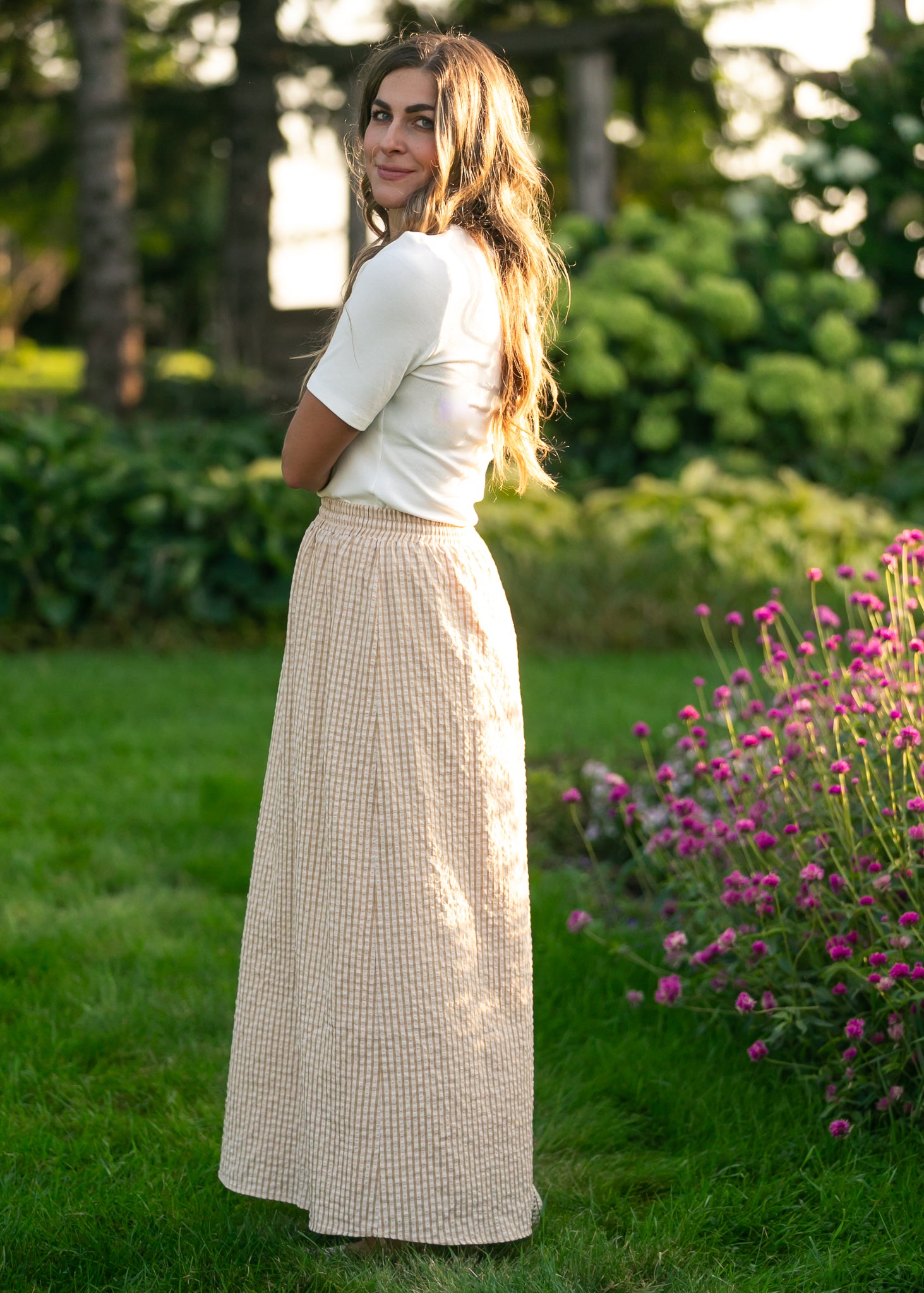 Woman in a white top and beige checkered skirt standing in a garden with greenery and flowers.