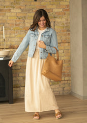 Woman in denim jacket and beige checkered dress holding a woven bag against a brick wall.