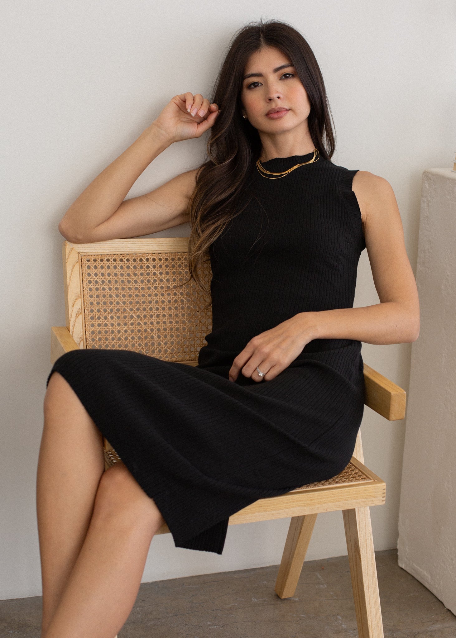 Woman in a black dress sitting on a wooden chair against a neutral wall.