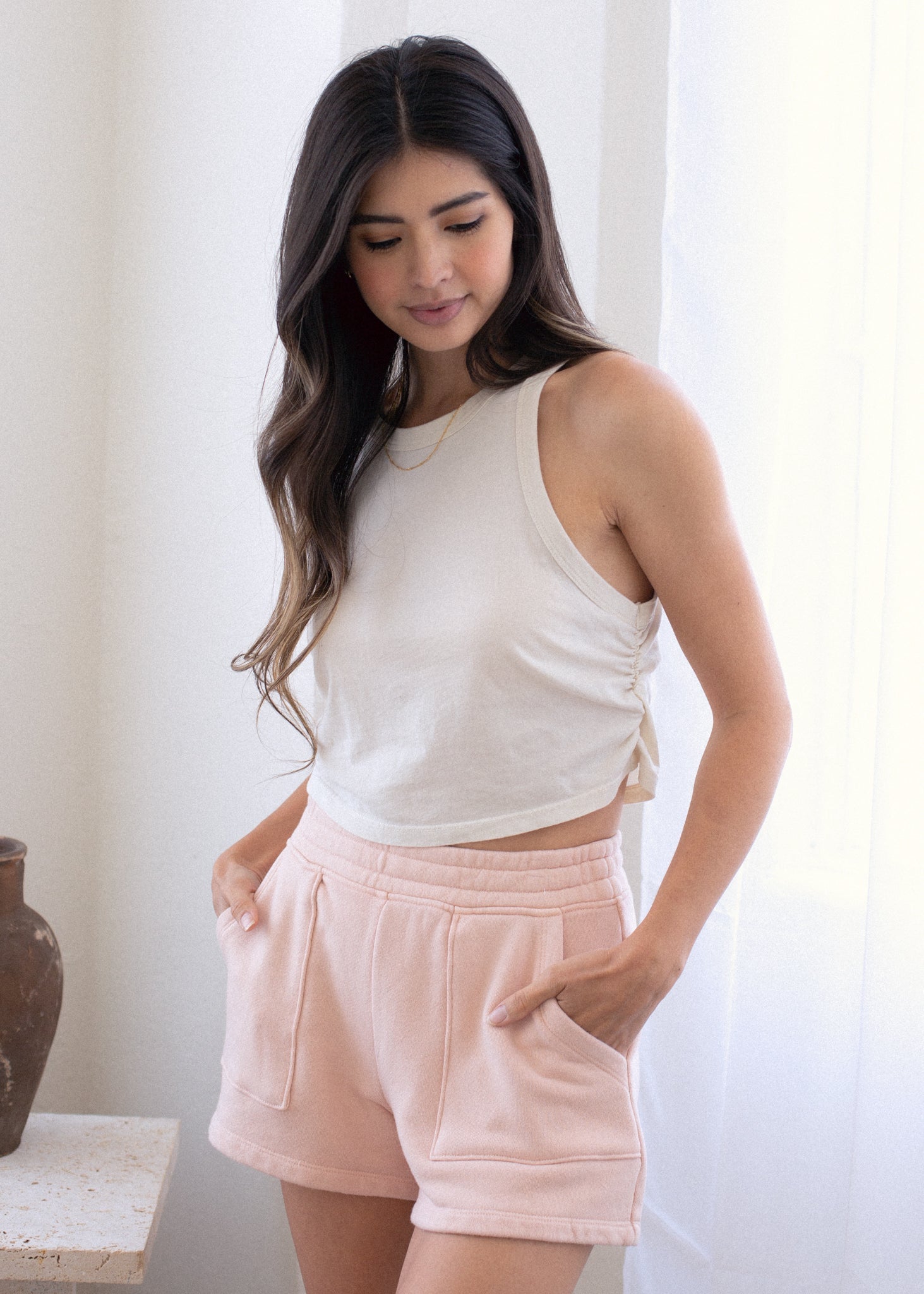 Woman wearing a white sleeveless top and pink shorts in a bright room.