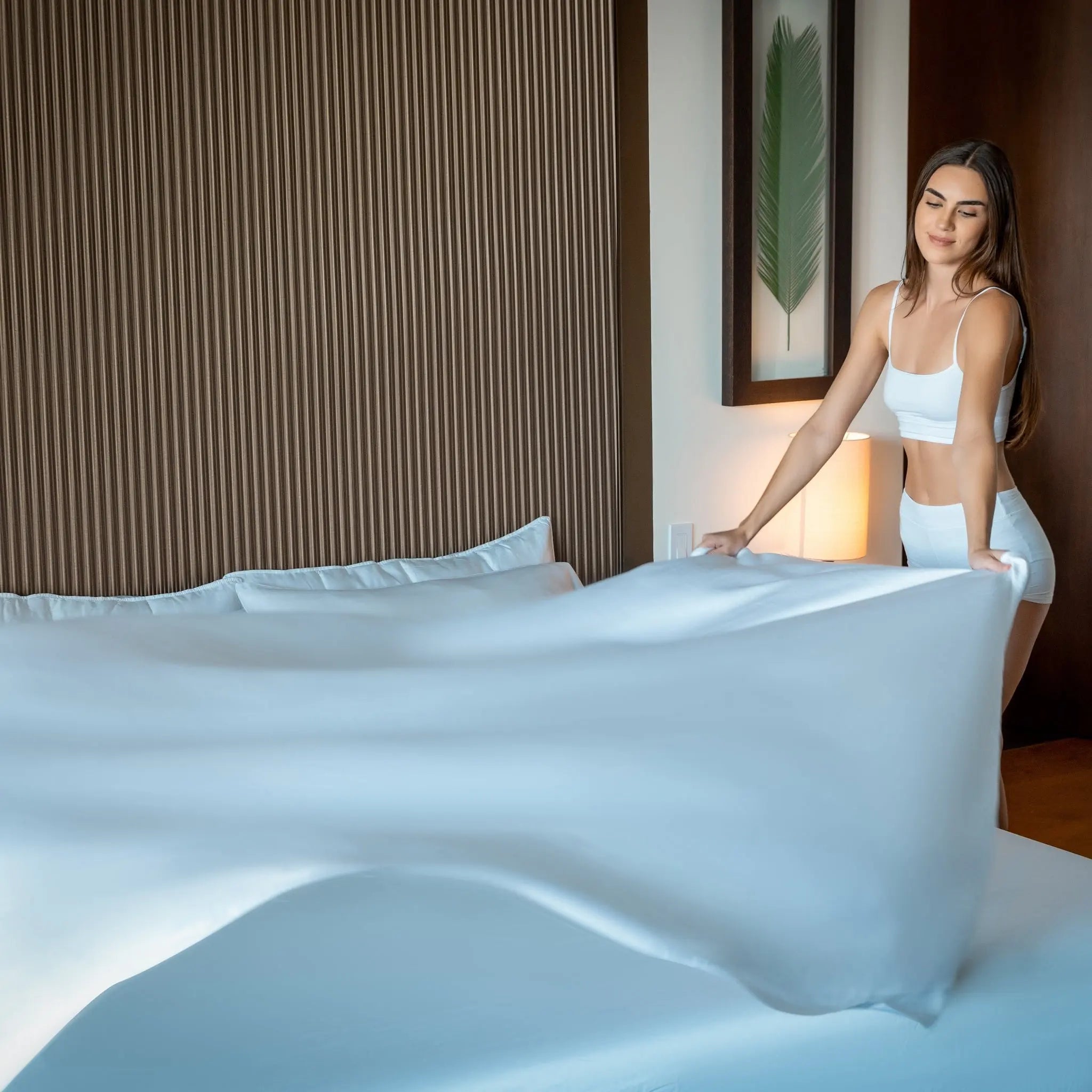 Woman in a white outfit arranging white bedding on a bed in a room with a lamp and wall art.