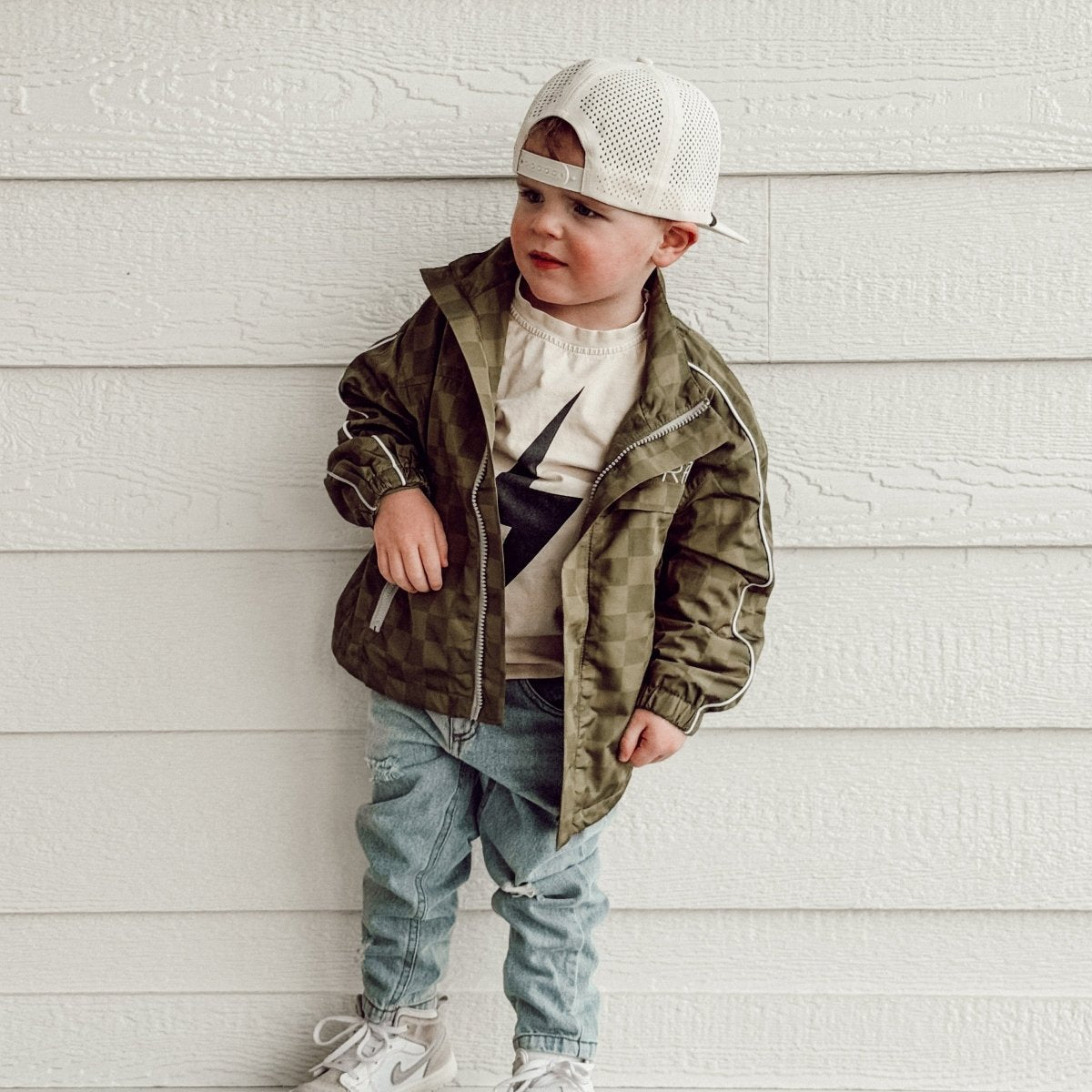 Child wearing a green jacket and white cap against a light wooden wall