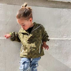 Child wearing a green camouflage jacket against a concrete wall