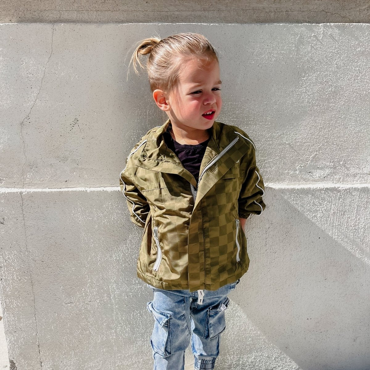 Child wearing a green jacket standing against a gray wall