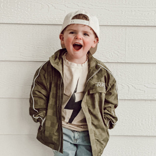 Child wearing a green jacket and white cap with a star design, standing against a light wooden wall.