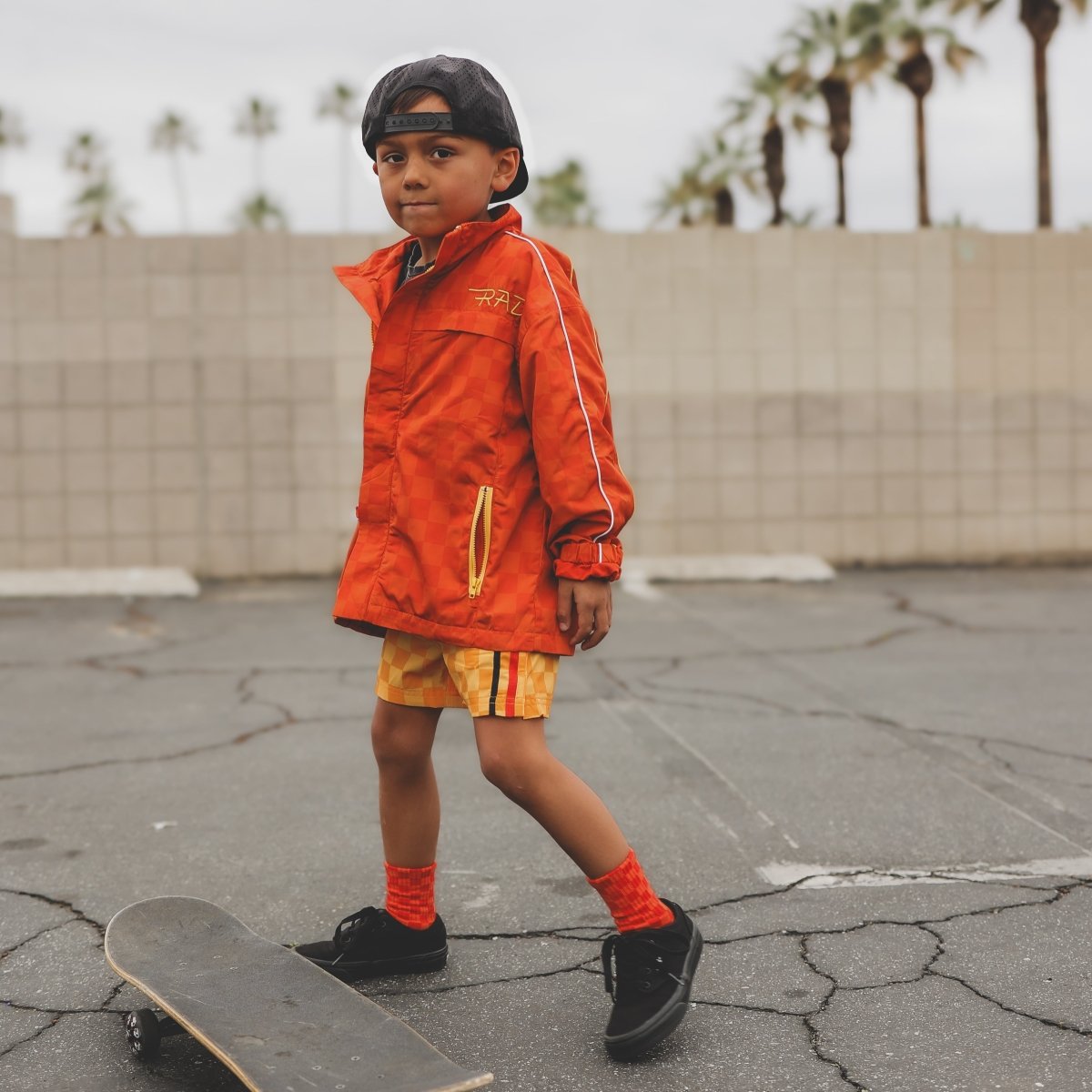 Child in an orange raincoat standing on a skateboard with palm trees in the background