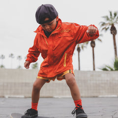 Child in an orange jacket with 'RAD' branding on a skateboard, palm trees in the background