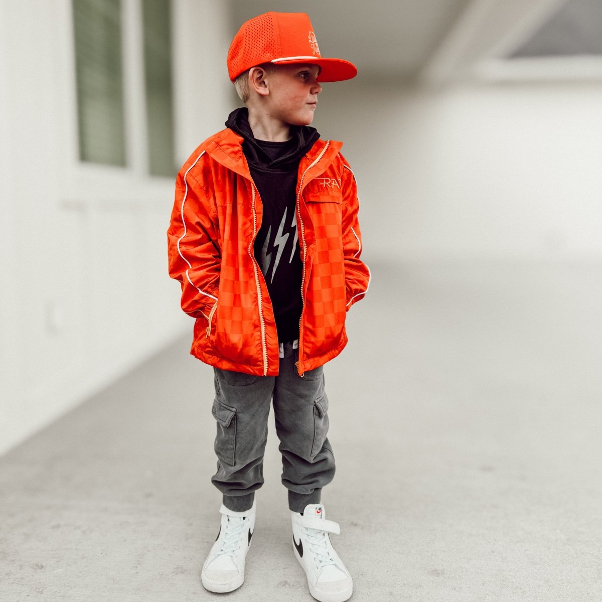 Child wearing an orange jacket, cap, and gray pants standing on a light-colored floor.