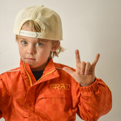 Child wearing an orange jacket with 'RAD' branding, beige cap, and making a hand gesture.